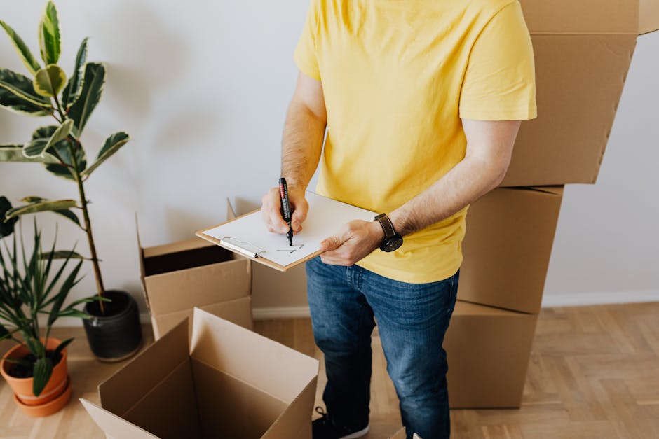 A person dressed in dark blue work clothing and wearing a headband stands inside a room with a textured light blue and white wall, surrounded by multiple cardboard moving boxes of various sizes, some